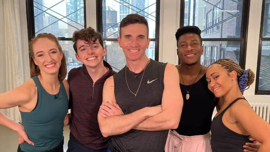 A group of five people smiling in front of a white wall with black windows.